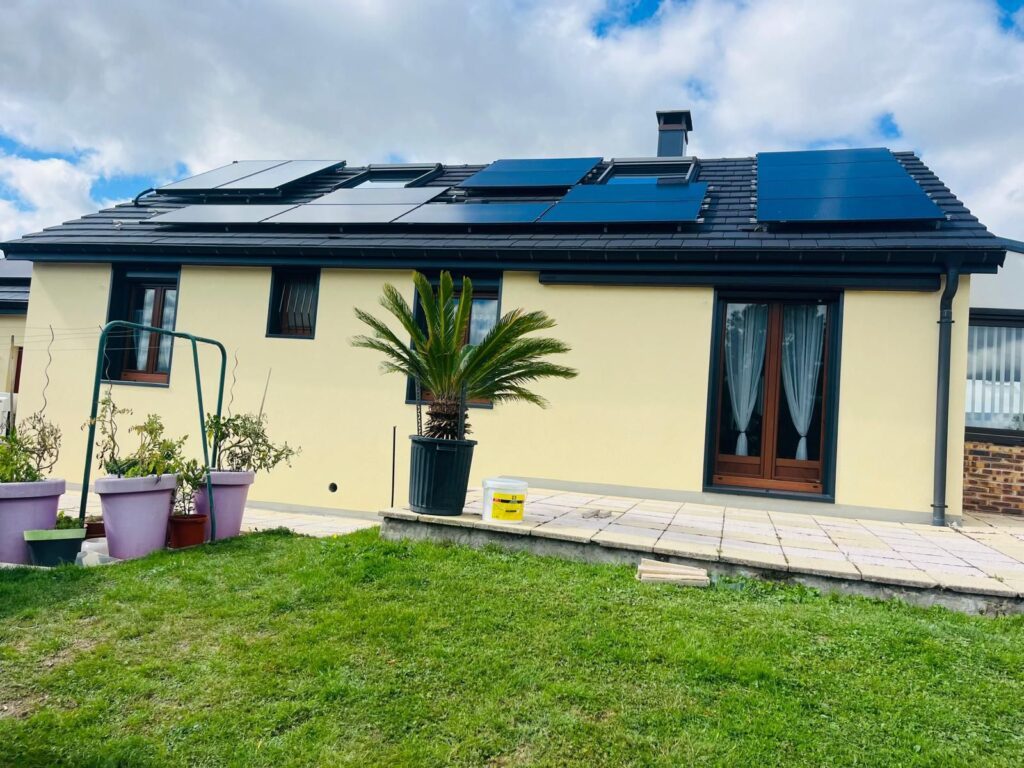 Maison rénovée avec façade jaune, toiture noire et panneaux solaires bleus, jardin paysagé avec palmier en pot