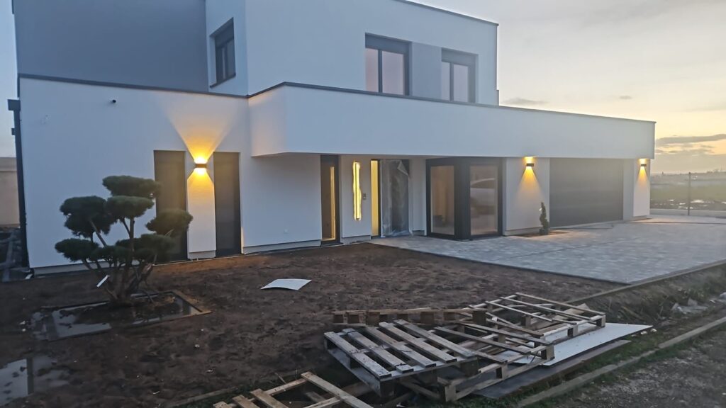 Maison moderne blanche en construction avec terrasse pavée, éclairage architectural et matériaux de chantier au sol